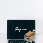 A laptop screen displaying 'Shop now' with a miniature shopping cart and cardboard box.